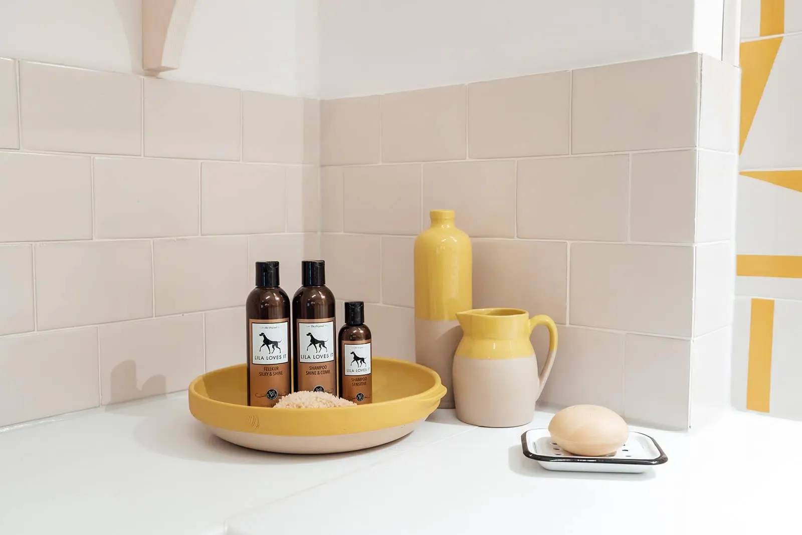 Drei braune Flaschen Hunde Fell Pflege Mittel stehen in einer gelben Waschschale mit Krügen und Seife in einem beige-weiß-gefließten Badezimmer | LILA LOVES IT "Fellkur Silky&Shine, Shampoo Shine&Comb & Shampoo Sensitive (Klein)"
