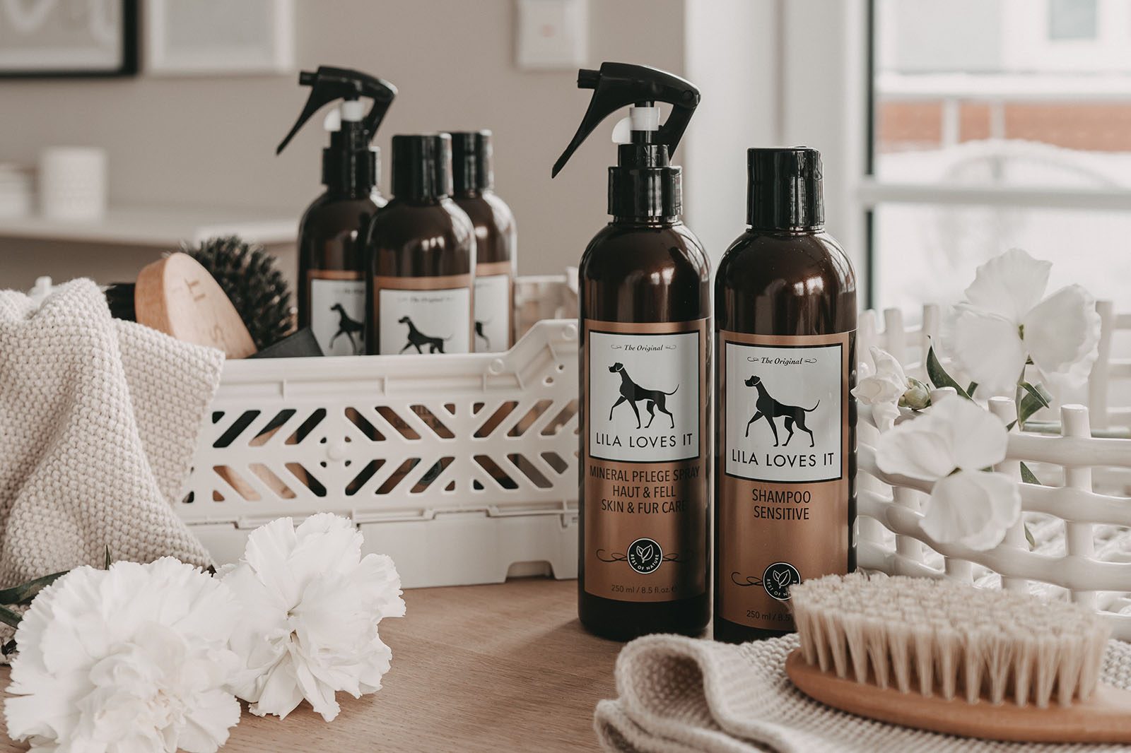 Braune Spüh- & Dosier-Flaschen mit einer Auswahl and unterschiedlichen Bürsten für Hundepflege auf einer braunen Holzoberfläche mit weißen Deko-Elementen | LILA LOVES IT "Mineral Pflege Spray Haut & Fell, Shampoo Sensitive & Welpenbürste"