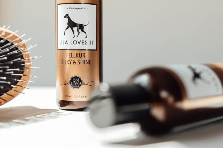 Dunkelbraune Shampooflaschen mit LILA LOVES IT Logo auf weißem Tisch mit angeschnittener Hundebürste links.
