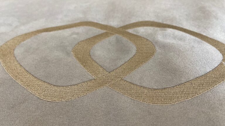 Ausschnitt einer individualisierten Stickerei auf einem grauen Hundebett mit einem goldenen Symbol von Verbindungsringen als Logo | LILA LOVES IT "Hotelbett für Hunde mit eigenem Logo"
