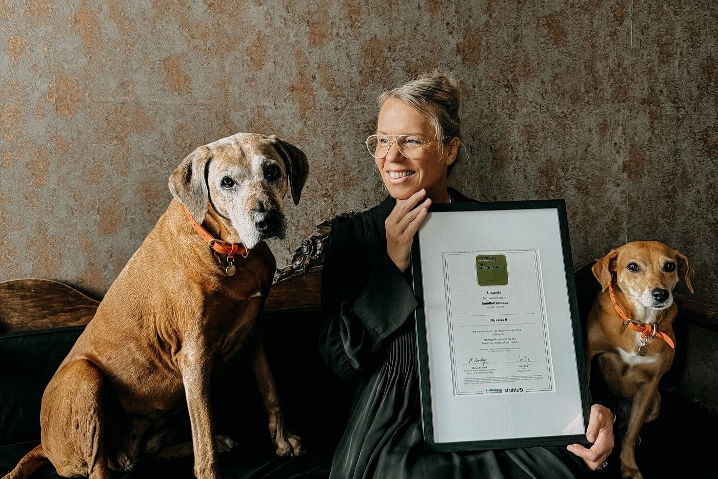 Gründerin Steffi Diem mit dem gerahmten Zertifikat für Lila loves it als Nummer 1 Marke für Fellpflege. Neben ihr sitzen ihre Hündinnen Lila und Penny.