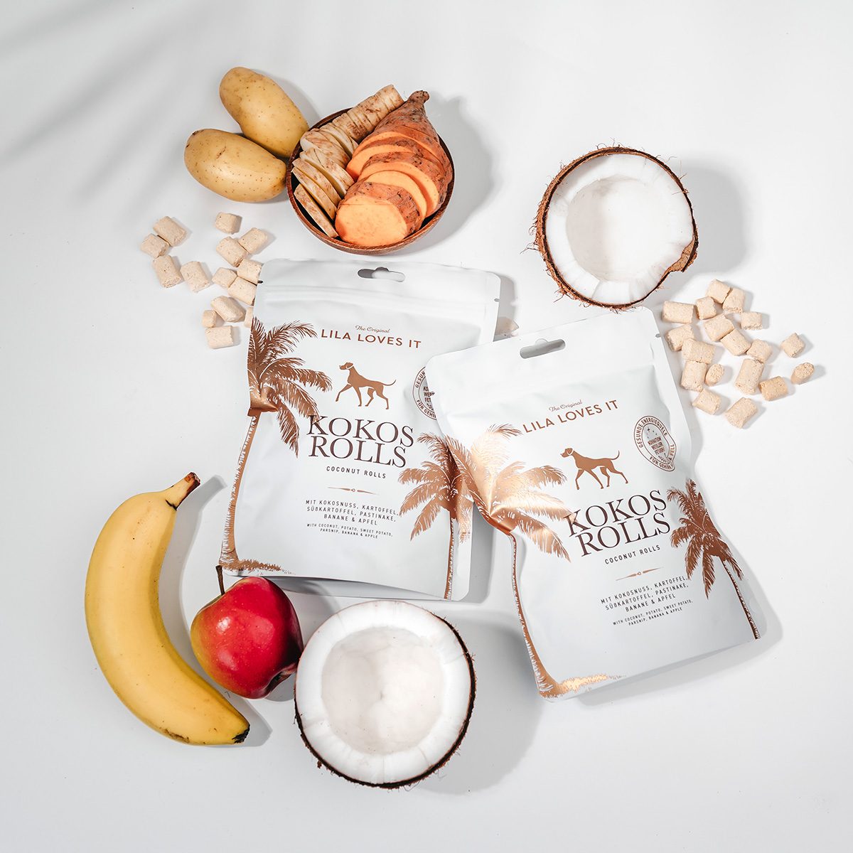Zwei Packungen LILA LOVES IT Kokos Rolls auf weißem Hintergrund, umgeben von Kokosnüssen, Banane, Apfel, Kartoffeln, Süßkartoffel und den hellen Hundesnacks.