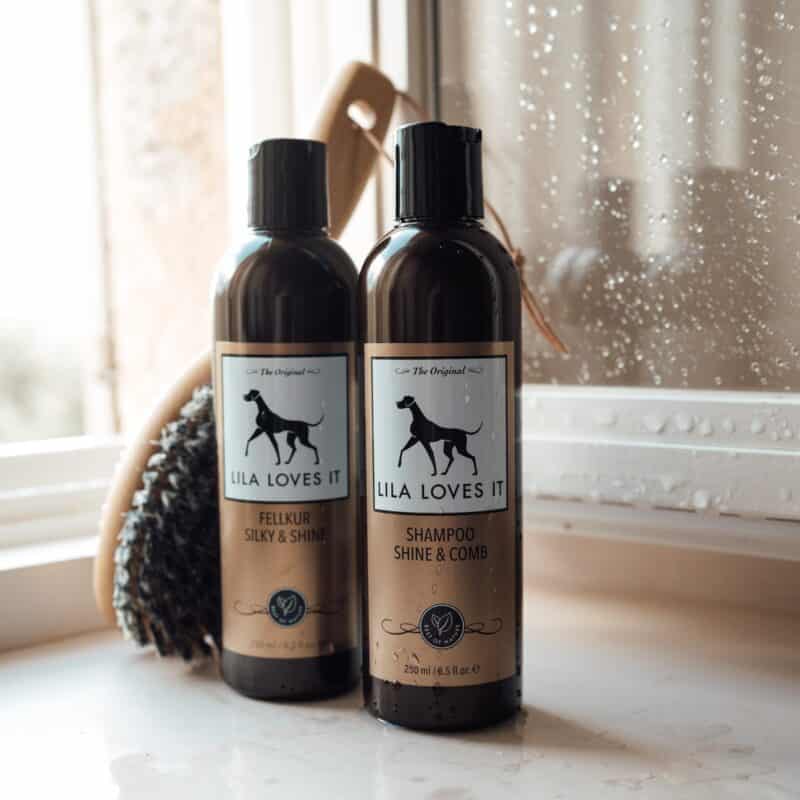 Zwei braune Flaschen mit Fellpflege für Hunde stehen neben einer Hundebürste mit Griff vor einem feuchten geöffneten Badezimmerfenster | LILA LOVES IT "Fellkur Silky & Shine, Shampoo Shine & Comb"
