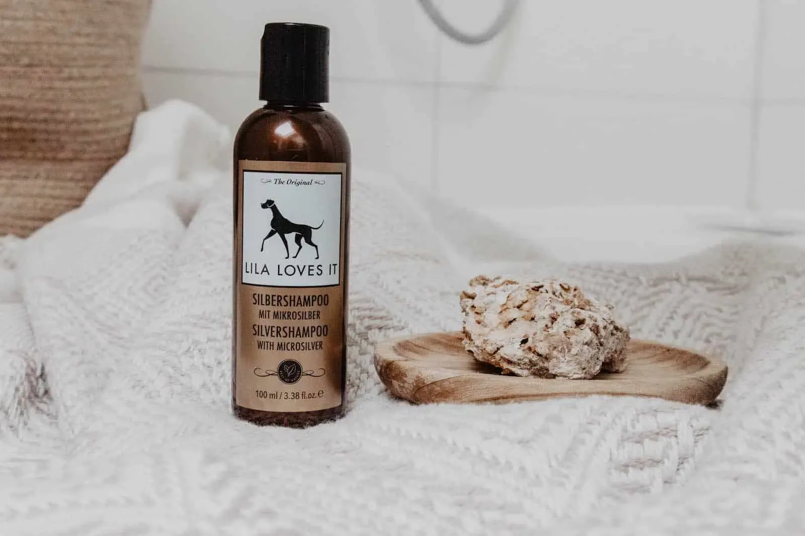 Kleine braune Flasche Shampoo für Hunde auf weißem Stickteppich neben einem Sandstein auf einer flachen Holzschale vor weißen Badezimmer Fliesen | LILA LOVES IT "Silbershampoo"
